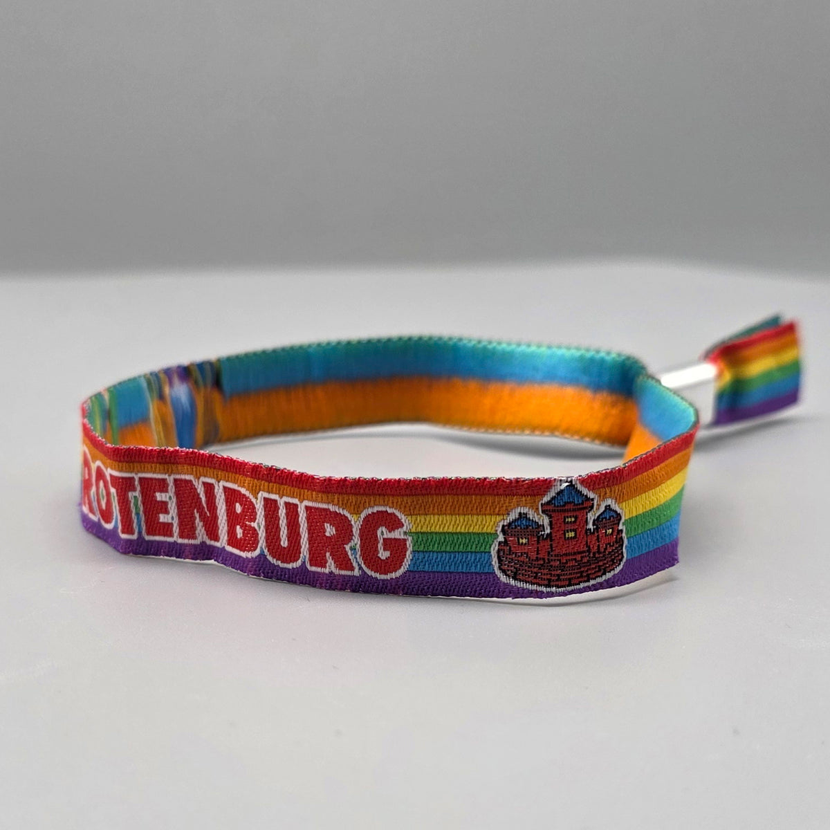Detailansicht des gewebten Stoffarmbands auf weißem Hintergrund. Auf dem farbintensiven Regenbogen-Muster leuchten der rote Schriftzug „ROTENBURG“ sowie ein klassisches rotes Burg-Wappenelement, das regionale queere Sichtbarkeit symbolisiert.