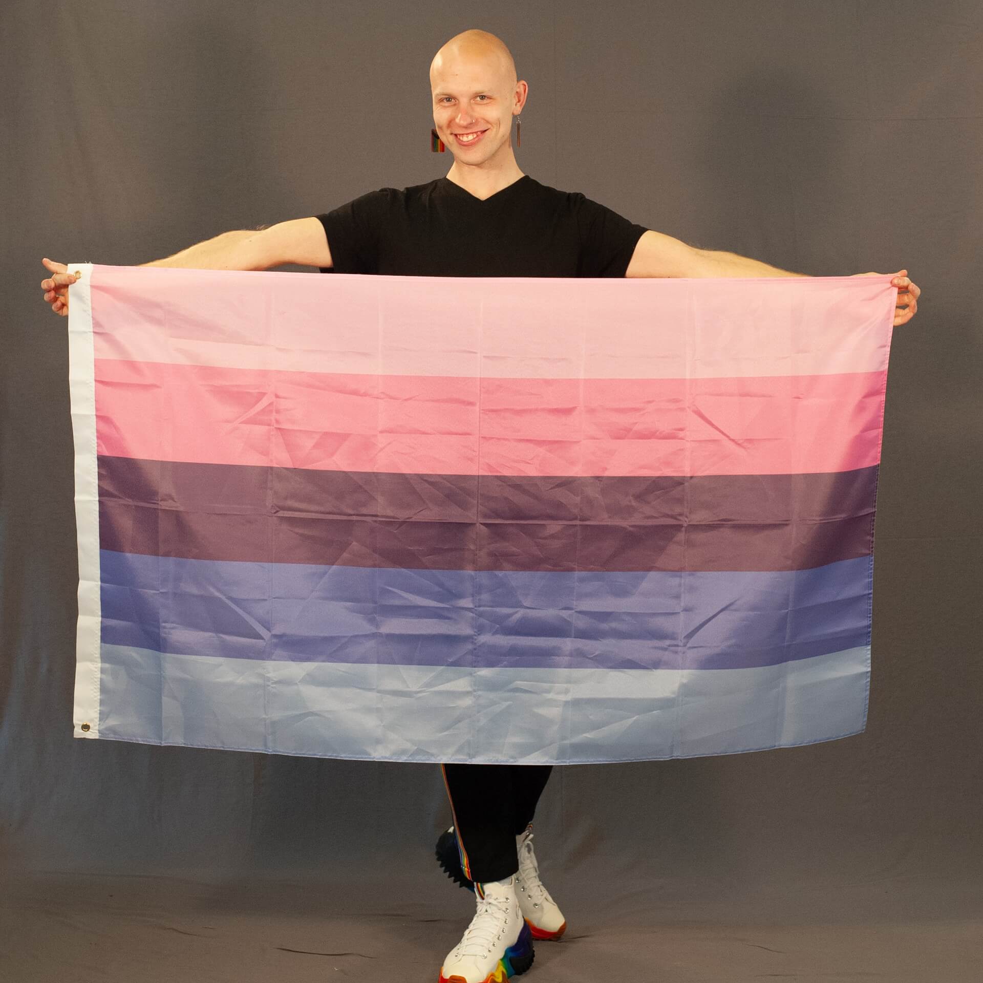 Ein Studiofoto des ehrenamtlichen Laienmodels Jonas LGBT, ein bekannter queerer Instagram-Aktivist, der die Omnisexuell Pride Flagge stolz präsentiert. Er hält die fünfstreifige Flagge mit den Farben Hellrosa, Pink, Dunkelblau, Blau und Hellblau mit beiden Armen weit ausgebreitet nach oben. Er blickt mit einem herzlichen Lächeln in die Kamera, trägt ein schwarzes V-Neck-T-Shirt, auffällige Regenbogen-Ohrringe und farbenfrohe Sneaker mit Plateausohle im Regenbogen-Design vor einem grauen Hintergrund.