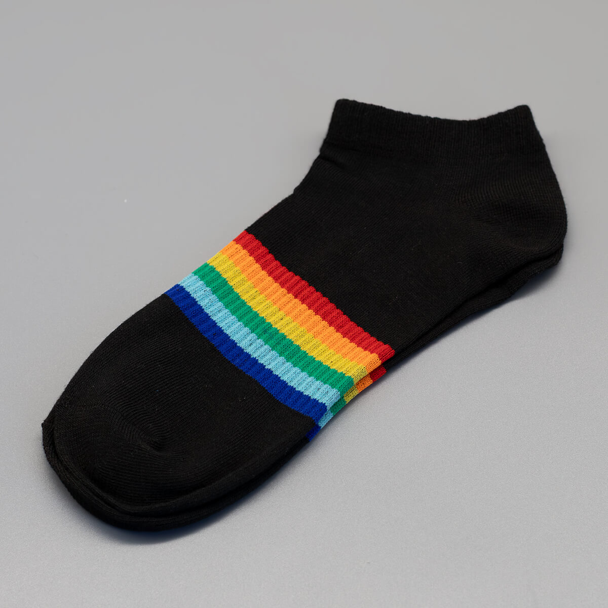 Ein freigestelltes Paar schwarzer Sneakersocken präsentiert sich mit einem horizontalen Streifenmuster direkt in der Mitte des Fußes. Die kräftigen Farben des klassischen LGBTIQ-Regenbogens setzen einen leuchtenden Kontrast auf dem dunklen Grundgewebe.