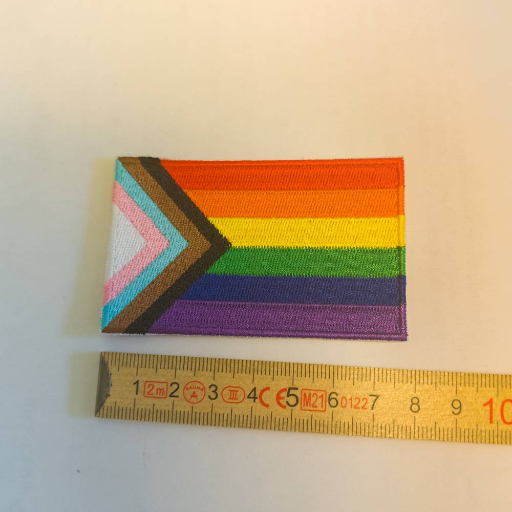 Ein gestickter Aufnäher der Progress Pride Flagge. Rechts die sechs Regenbogenstreifen, von links ragt der Keil in Weiß, Rosa, Hellblau, Braun und Schwarz ins Bild. Der Patch liegt auf einem neutralen Hintergrund neben einem gelben Holz-Zollstock, der die Maße des Aufnähers verdeutlicht.
