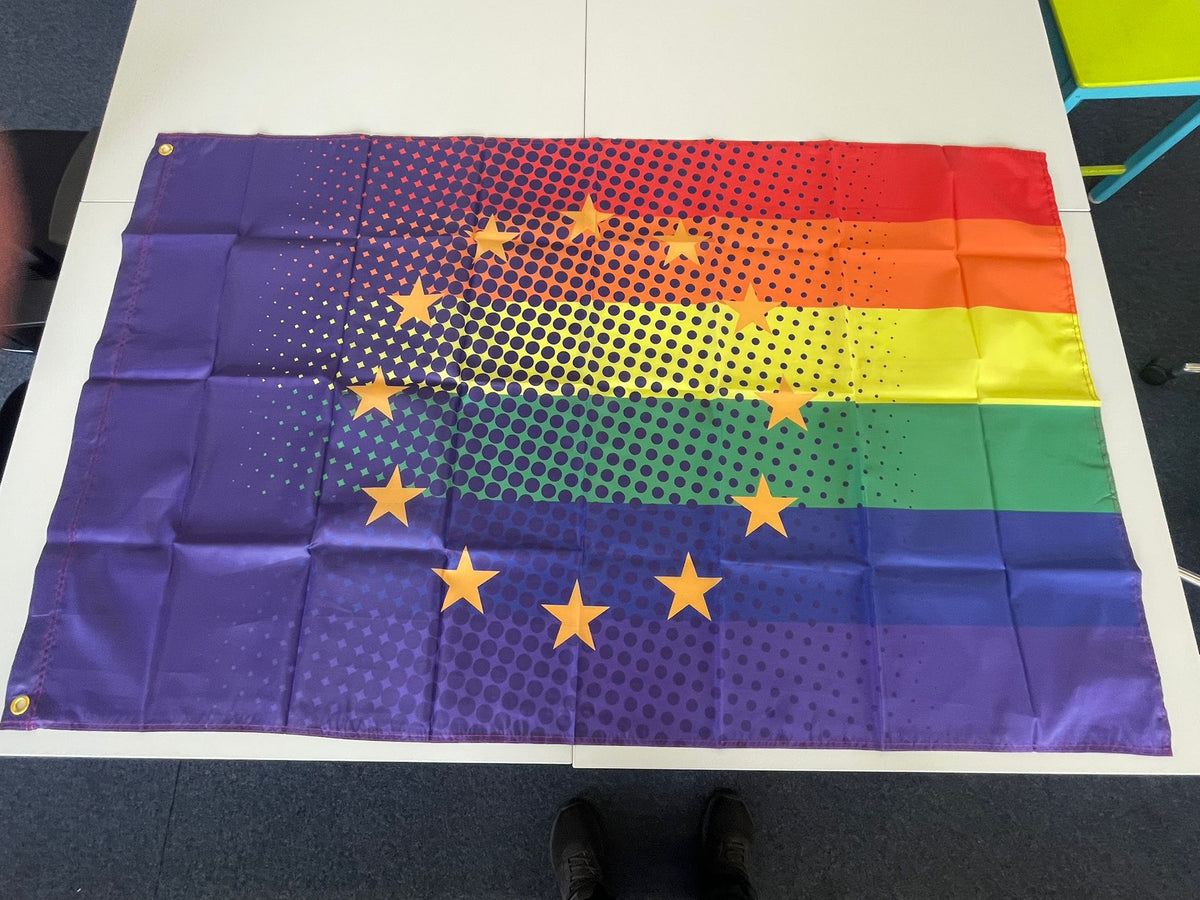 Die europäische Pride-Flagge liegt flach ausgebreitet auf zusammengestellten weißen Tischen und zeigt das vollständige, ungelabelte Design. Gut zu erkennen sind das kräftige Farbschema der LGBTIQ-Bewegung, der gelbe Sternenkreis der EU sowie die beiden goldfarbenen Metallösen an der verstärkten linken Kante.