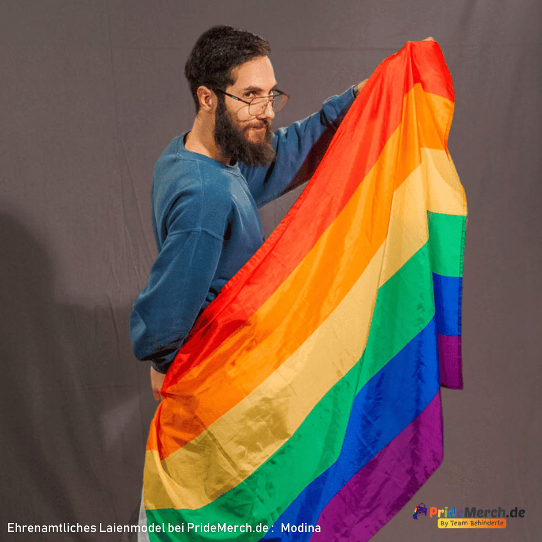Laienmodell Modina steht vor einem grauen Hintergrund und hält die Regenbogenflagge mit seiner linken Hand nah an sein Gesicht nach oben. Er blickt leicht über den Rand seiner Brille hinweg nachdenklich zur Seite. Die starken Farben der Flagge bilden einen deutlichen Kontrast zu seinem dunkelblauen Pullover.