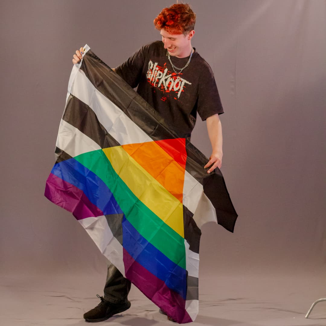 Das Laienmodell Justus blickt mit einem breiten Lächeln schräg nach unten. Er trägt ein schwarzes Band-Shirt und dunkle Hosen. Er hält die Straight Ally Flagge so vor seinem Körper, dass der Stoff leicht in Falten fällt. Das leuchtende, regenbogenfarbene „A“ auf dem schwarz-weißen Streifenhintergrund sticht deutlich als zentrales Motiv hervor.