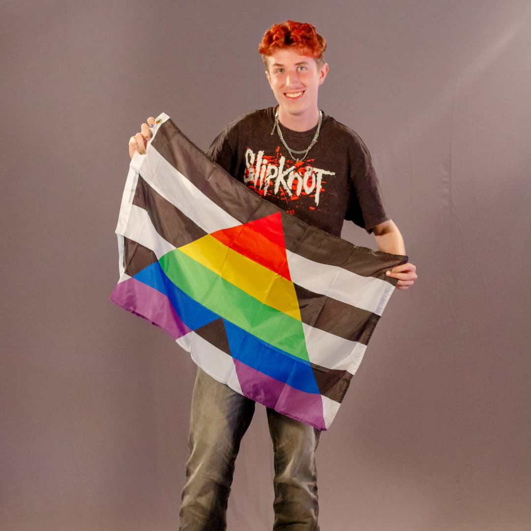 Das ehrenamtliche Laienmodell Justus Kornmesser steht lachend vor einem grauen Fotohintergrund. Er hat kurze, rötlich gefärbte Haare, trägt ein schwarzes „Slipknot“-T-Shirt, mehrere silberne Halsketten und eine dunkelgraue Jeans. Mit beiden Händen hält er die schwarz-weiße Straight Ally Flagge mit dem bunten Regenbogen-A vor sich aufgefaltet.