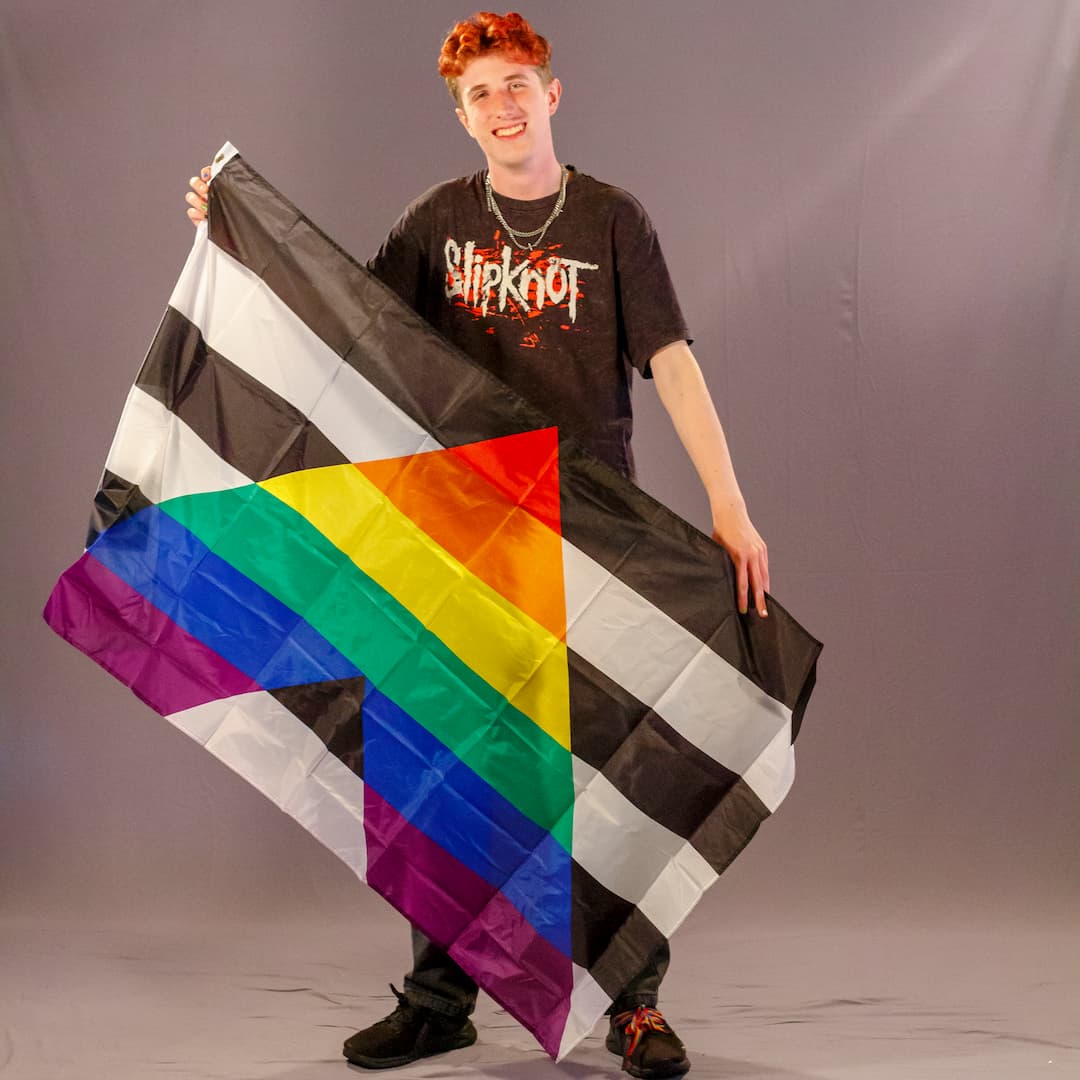 Justus Kornmesser, ein Laienmodell mit rötlichen Haaren und schwarzen Sneakern mit bunten Schnürsenkeln, blickt direkt und freudig in die Kamera. Er breitet die Straight Ally Flagge großflächig vor seinen Beinen und seinem Oberkörper aus. Das kontrastreiche Design – schwarz-weiße Querstreifen durchbrochen von einem großen Regenbogen-Dreieck – ist in seiner vollen Breite gut zu erkennen.