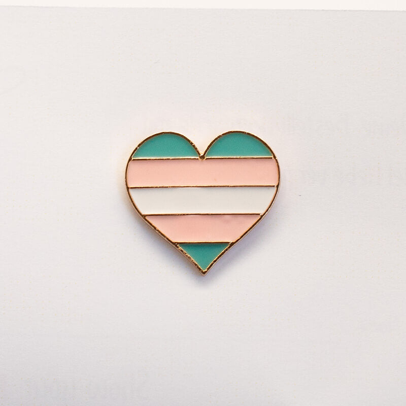 Die hellblauen, rosa und weißen Streifen der Trans-Community zieren diese glänzende Anstecknadel im Herzdesign. Das Produktbild fokussiert die glatte Soft-Emaille-Oberfläche und das kontrastierende Metallelement vor dem neutralen Studiohintergrund.