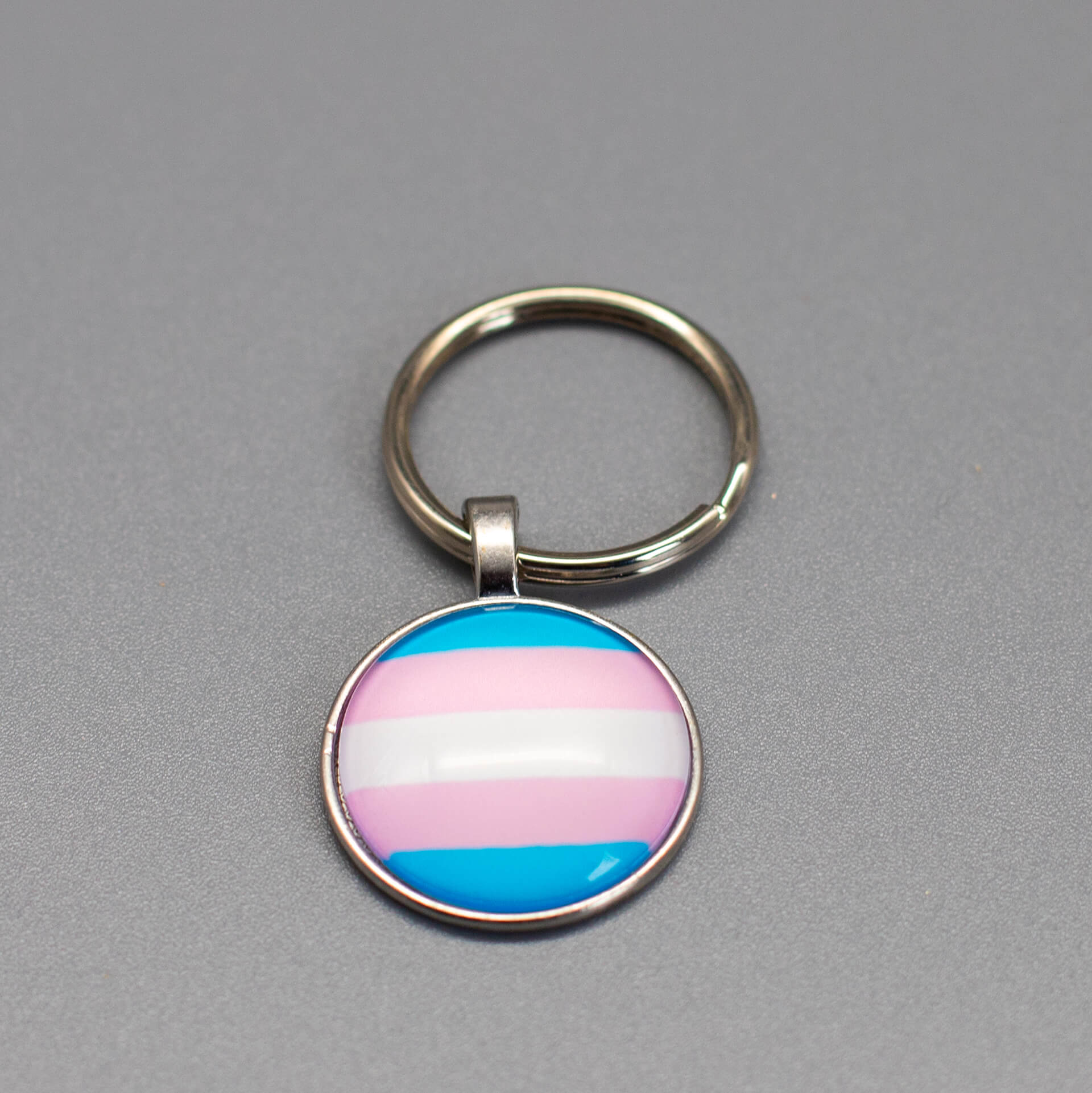 Runder LGBTQ Transgender Schlüsselanhänger in Silber mit gewölbtem Glas und Trans Pride Flagge auf grauem Hintergrund