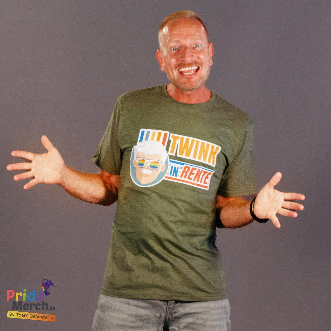„Mann mit lächelndem Gesicht und ausgestreckten Armen trägt ein olivgrünes T-Shirt mit dem PrideLab-Design 'Twink in Rente'. Das Design zeigt eine stilisierte Abbildung eines älteren Mannes mit weißem Haar und Regenbogen-Sonnenbrille sowie den Text 'TWINK IN RENTE' in bunten, lebendigen Farben. Das Shirt wird von pridemerch.mom präsentiert – einem Online-Shop für LGBTQ+-Merchandise, der von Team Behinderte betrieben wird. Der humorvolle und stolze Look verbindet Pride-Kultur mit spielerischer Selbstironie.
