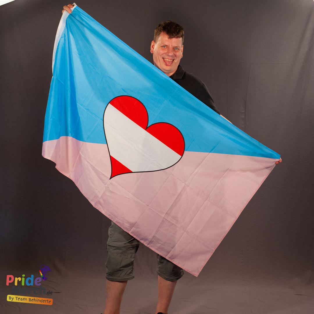 In der originalen Studioaufnahme präsentiert das ehrenamtliche Laienmodel Rene gut gelaunt die weit geöffnete Ageplay Pride Flagge vor einem dunkelgrauen Hintergrund. Er hält die große Fahne im Format 150x90 cm mit beiden Händen ausgebreitet, sodass das diagonale Farbdesign in Hellblau und Rosa mit dem zentralen rot-weiß-roten Herz-Symbol perfekt sichtbar wird. Unten links im Bild ist das Logo von pridemerch.mom (By Team Behinderte) eingebunden.