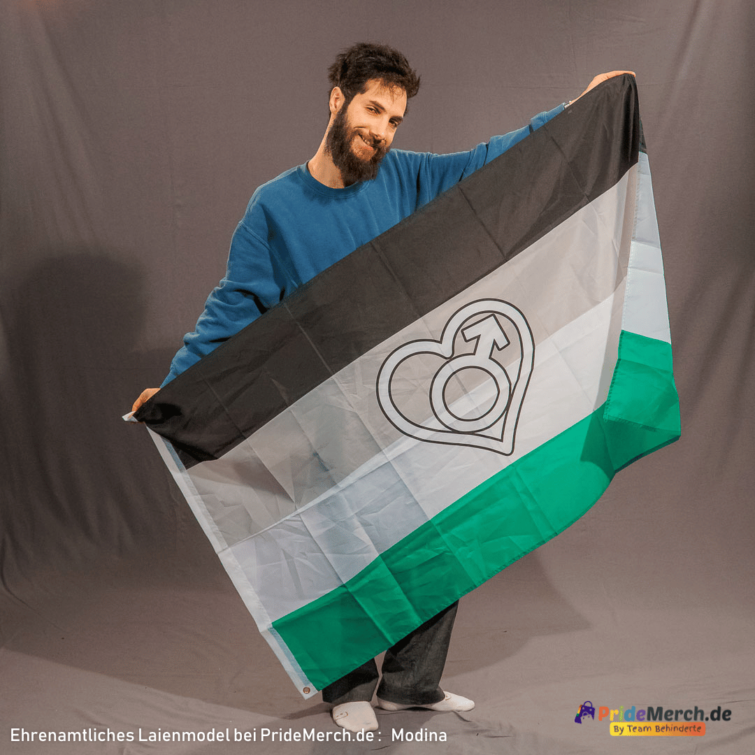 In der Studioaufnahme präsentiert das ehrenamtliche Laienmodel Modina mit einem freundlichen Lächeln und vollem Bart die geöffnete Androphilie Pride Flagge vor grauem Studiohintergrund. Er hält die Pride Flagge im Format 150x90 cm schräg ausgebreitet, sodass die vier Querstreifen in Schwarz, Grau, Weiß und Grün sowie das markante weiße Herz mit dem Mars-Symbol im Zentrum perfekt zu sehen sind. Unten rechts befindet sich das Logo von pridemerch.mom.