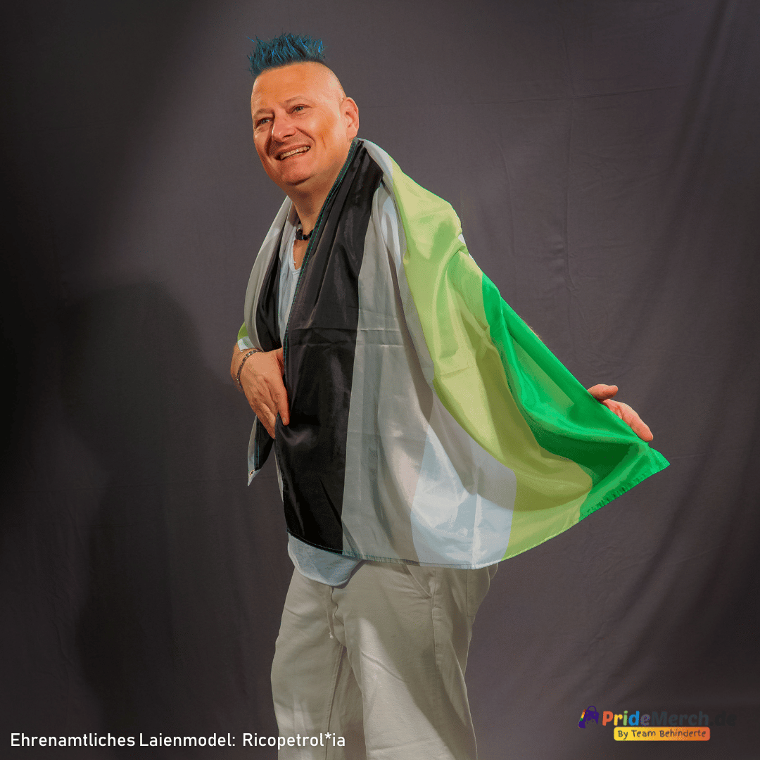 Das ehrenamtliche Laienmodel Ricopetrolia (bekannt als Bremer Drag Queen Gina Petrolia) präsentiert gut gelaunt im Profil die große Aromantische Pride Flagge im Format 150x90 cm. Das Foto zeigt das Model mit blau gefärbtem Irokesenschnitt, wie es sich die Flagge mit den Farben Dunkelgrün, Hellgrün, Weiß, Grau und Schwarz stolz wie einen Umhang über die Schulter gelegt hat. Unten rechts ist das Logo von pridemerch.mom zu sehen.