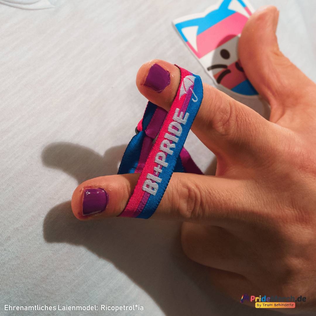 Porträt des ehrenamtlichen Laienmodels Gina Petrolia, die mit einem charmanten Lächeln für pridemerch.mom posiert. Sie blickt in die Kamera und hält ihre Hand an die Wange, wodurch das pink-lila-blaue Bi+ Pride Stoffarmbändchen an ihrem Handgelenk perfekt präsentiert wird. Unten links ist der Schriftzug „Ehrenamtliches Laienmodel: Gina Petrolia“ eingeblendet, rechts das Logo von pridemerch.mom – By Team Behinderte.