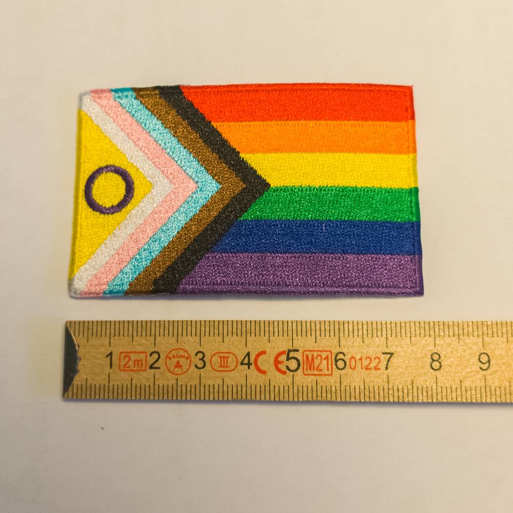 Ein strukturierter, gestickter Aufnäher der Intersex Inclusive Progress Flagge. Neben dem mehrfarbigen Keil befindet sich ganz links das gelbe Dreieck mit dem violetten Kreis der Intersex-Community. Der Aufnäher ist neben einem aufgeklappten gelben Zollstock platziert, der die exakten Abmessungen zeigt.