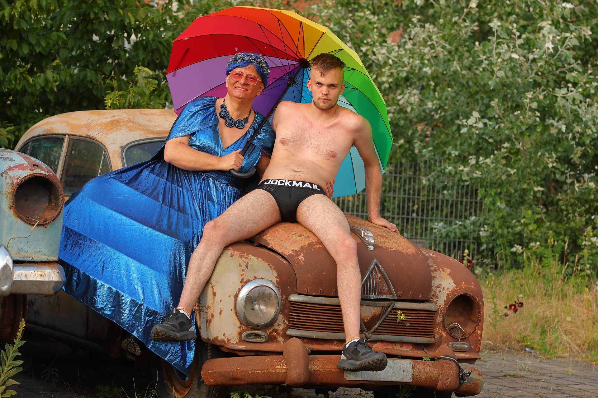 Drag Queen Gina Petrolia im metallisch blauen Kleid und Model Leo in schwarzer Jockmail-Unterwäsche posieren selbstbewusst auf der Motorhaube eines rostigen Oldtimers. Unter einem aufgespannten Regenbogen-Schirm blicken beide direkt in die Kamera – ein geheimnisvoller visueller Teaser für das, was bald kommt.